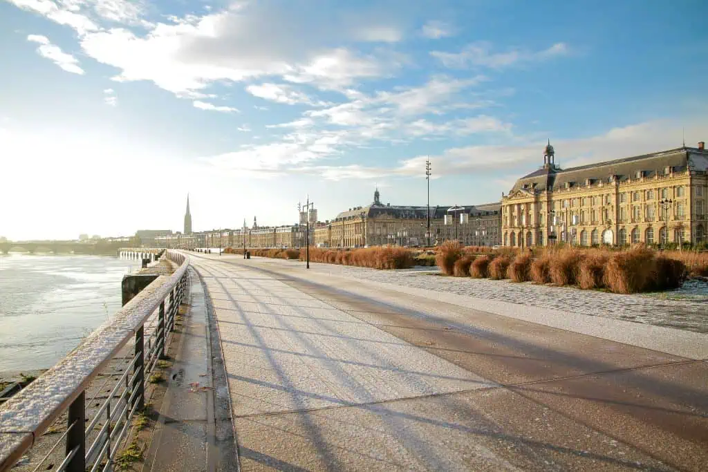 A picturesque riverside promenade in a city, with elegant historic buildings lining the right side. The walkway is lightly dusted with snow, and the sky is clear with scattered clouds, allowing the bright sunlight to cast long shadows. A church spire and a bridge are visible in the distance, adding to the charm of the urban landscape. The calm river runs along the left side, bordered by a railing. The scene is serene and peaceful, with a blend of natural and architectural beauty.
